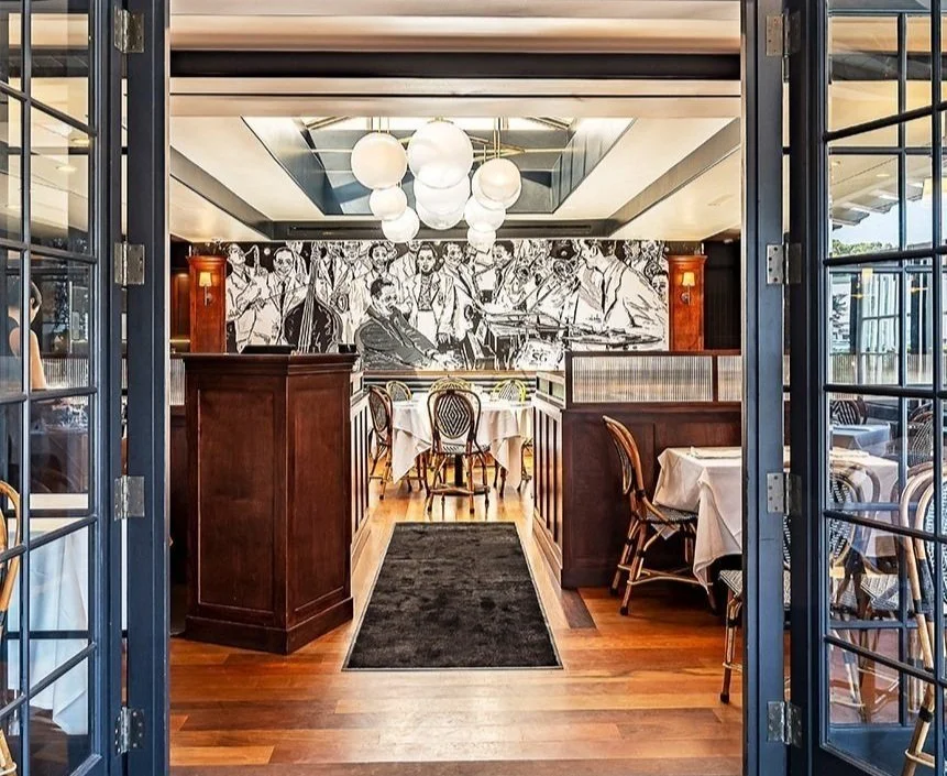 Interior of a restaurant with wooden flooring, dark wood partitions, tables with white tablecloths, a large mural on the wall, and hanging spherical lights.