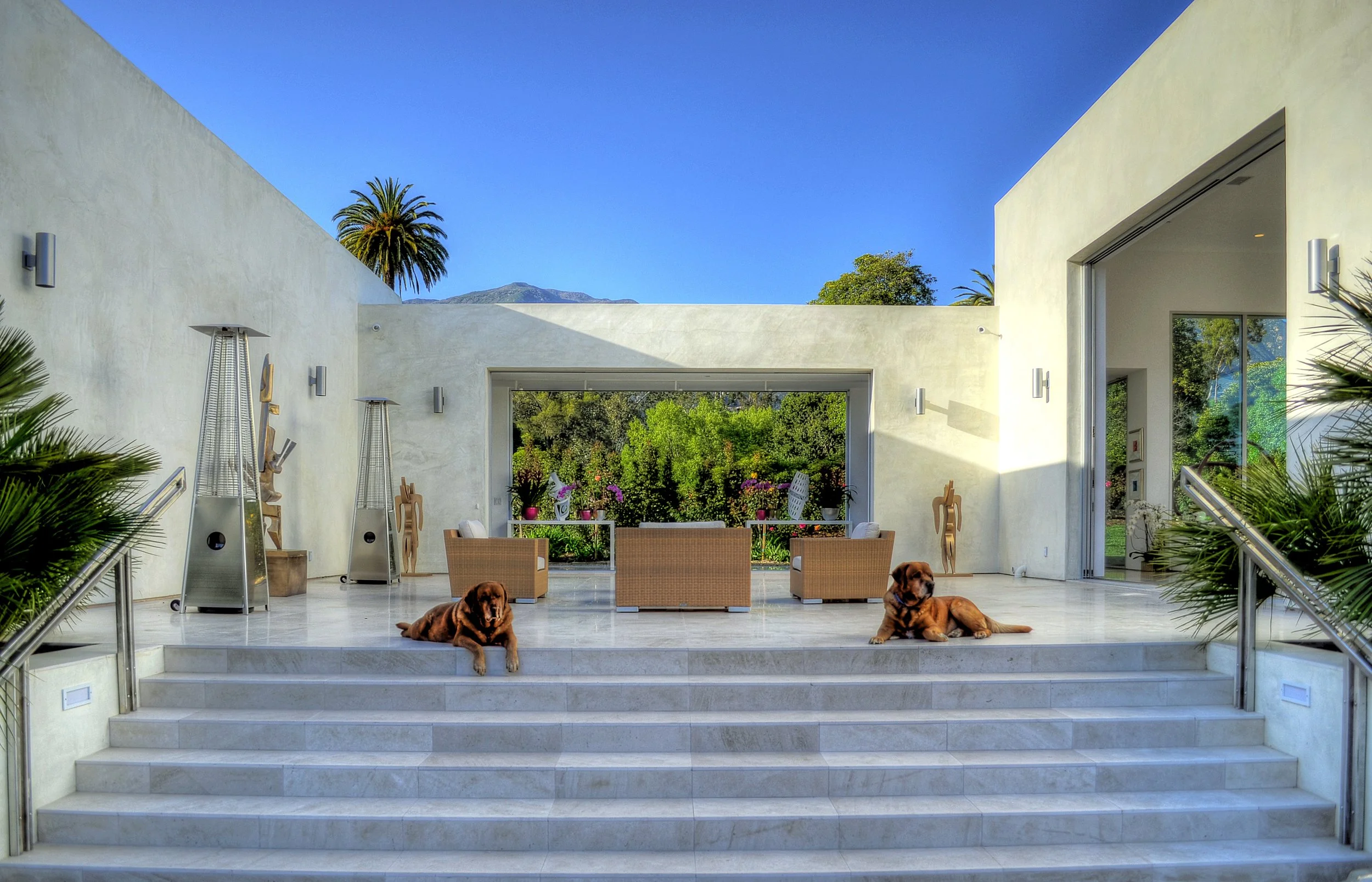 Modernist outdoor seating area for the Demetrios Residence, emphasizing symmetry and openness. Two dogs rest on the marble stairs, complementing the serene backdrop of lush greenery and mountain views.