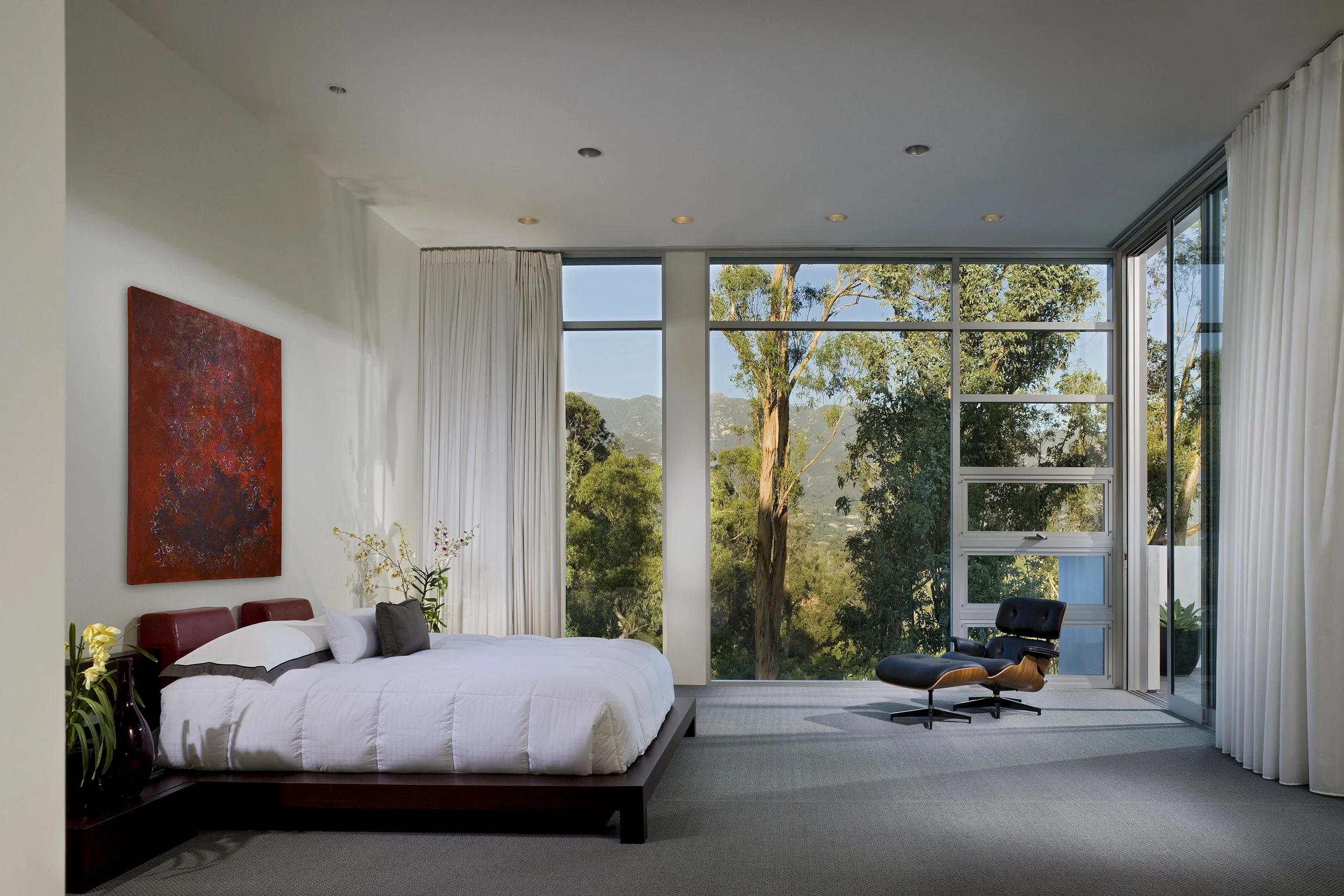 Modern bedroom with large floor-to-ceiling windows offering scenic mountain and tree views, featuring minimalist decor and a comfortable seating area.