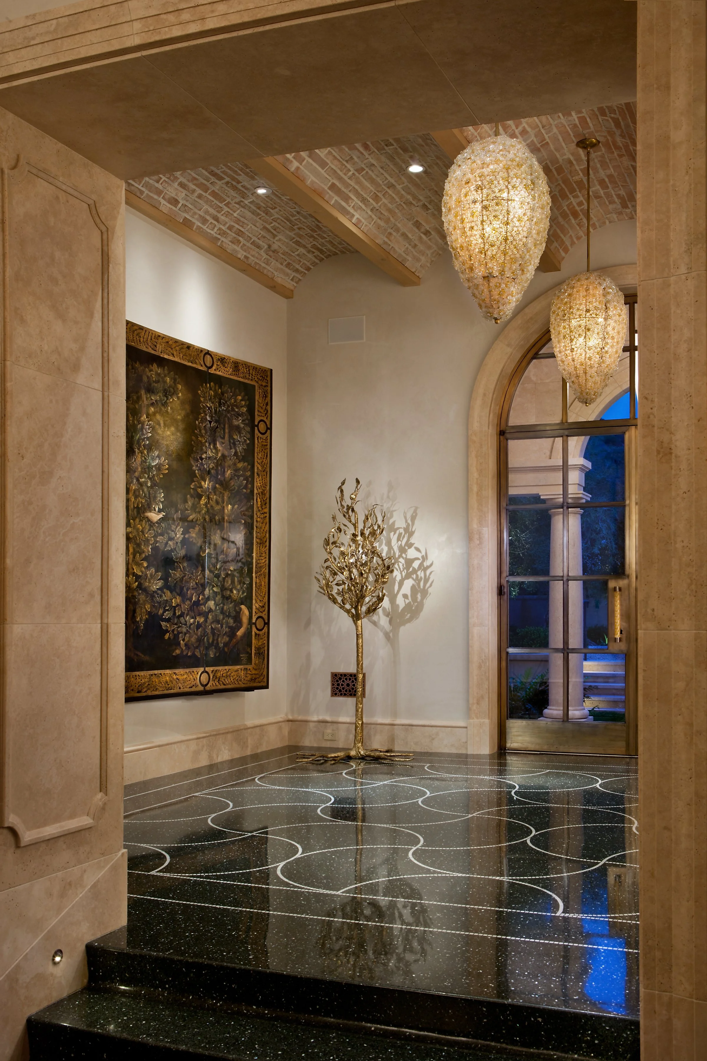 Elegant foyer with decorative lighting, marble floors, arched window, ornate painting, and metallic tree sculpture.