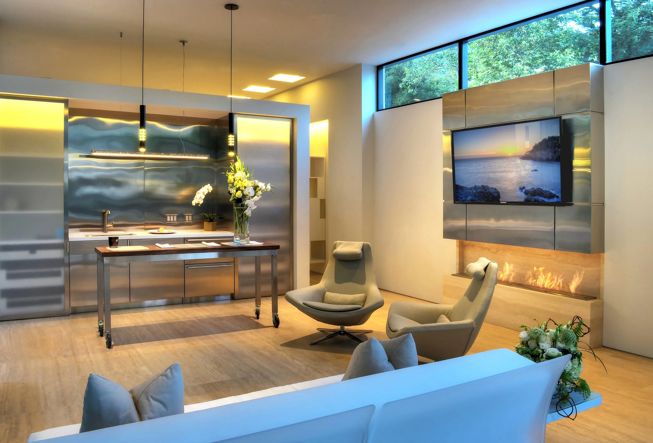 Contemporary living space featuring a sleek stainless steel kitchen, minimalist lounge chairs, and a fireplace below a wall-mounted television, designed by The Warner Group Architects, Inc.
