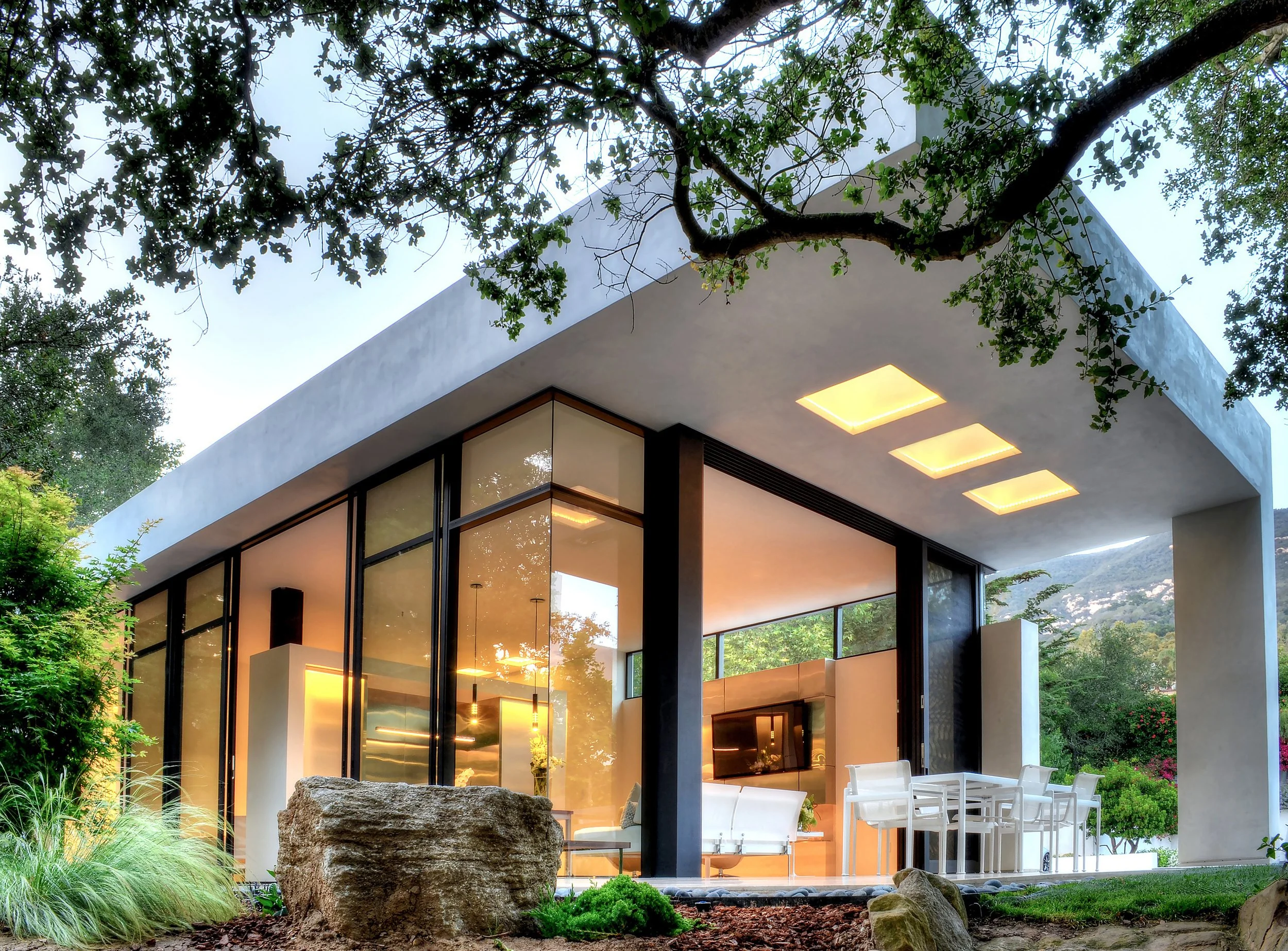 Modern architectural home with floor-to-ceiling windows, illuminated ceiling lights, and outdoor dining space, surrounded by natural landscaping, designed by The Warner Group Architects, Inc.