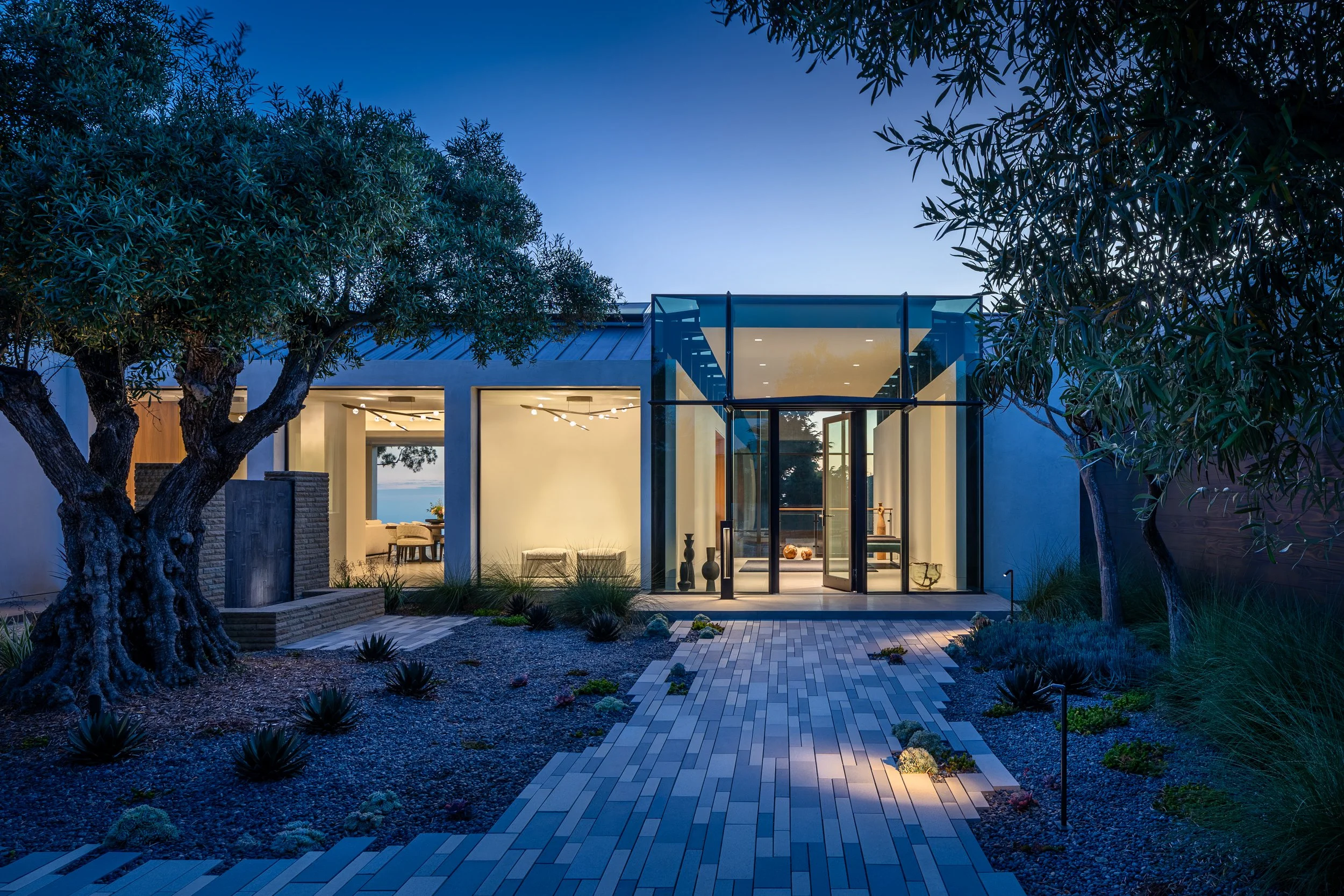 Modern glass-front house with illuminated interior and landscaped garden at dusk.