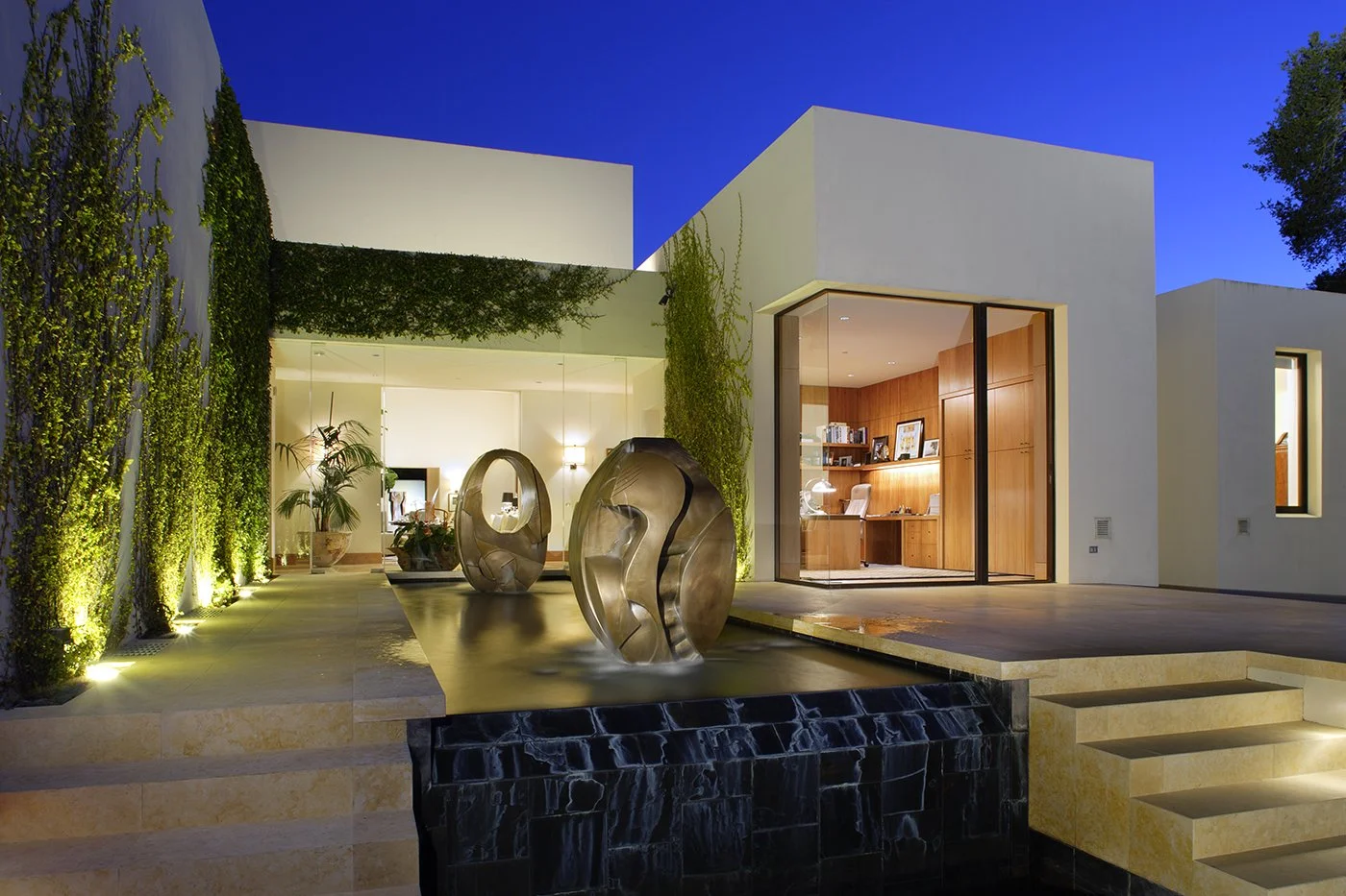 A modern architectural entryway featuring illuminated greenery and artistic sculptures leading into the home office.