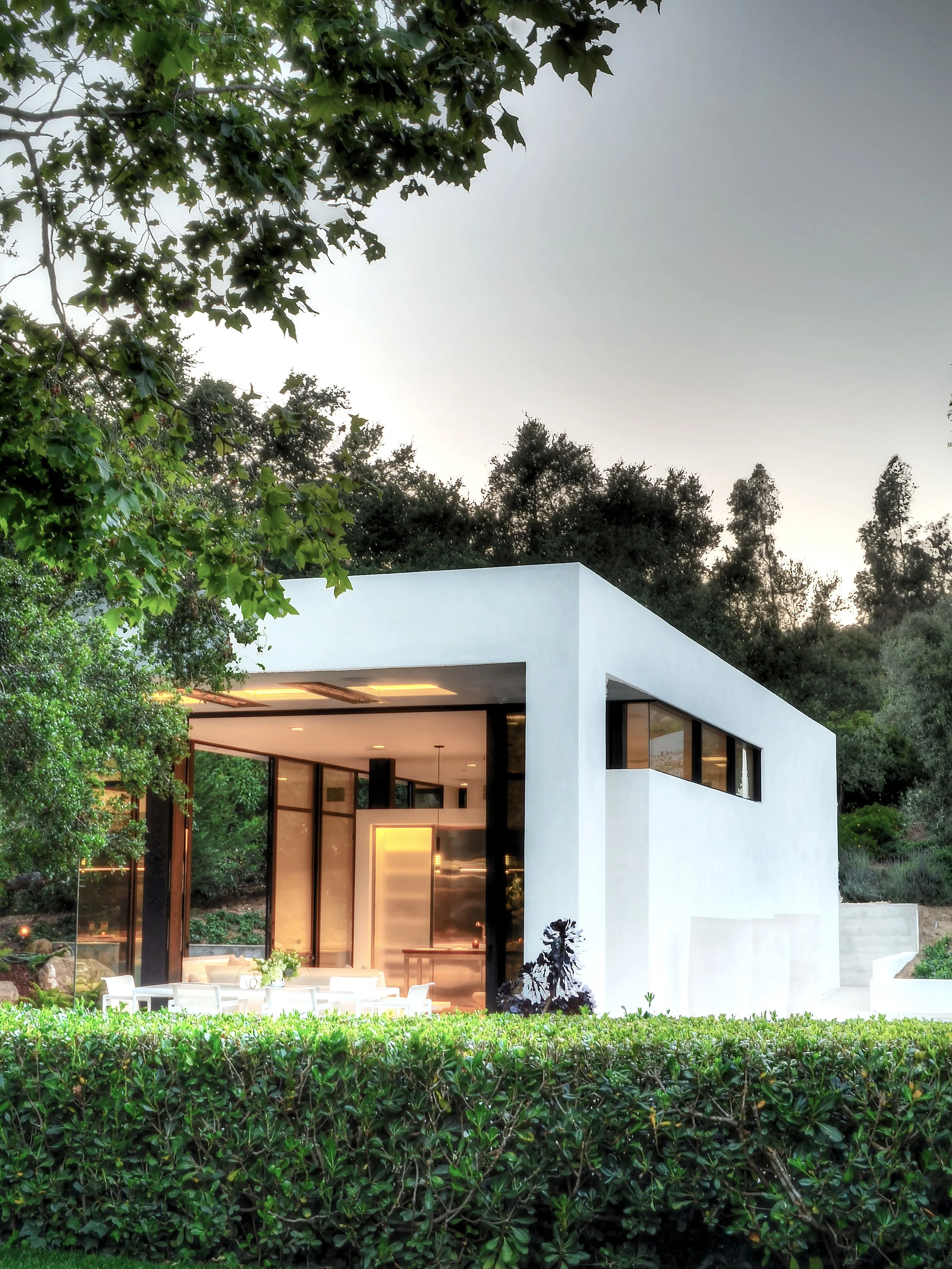 Modern white structure surrounded by lush greenery, featuring expansive glass windows and an outdoor dining area, designed by The Warner Group Architects, Inc.