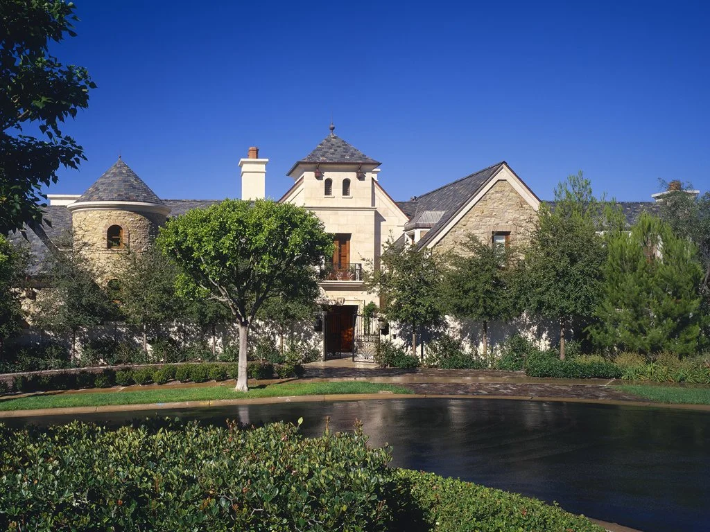 Large stone house with a round turret, steeply pitched roofs, and surrounded by manicured landscaping and trees.