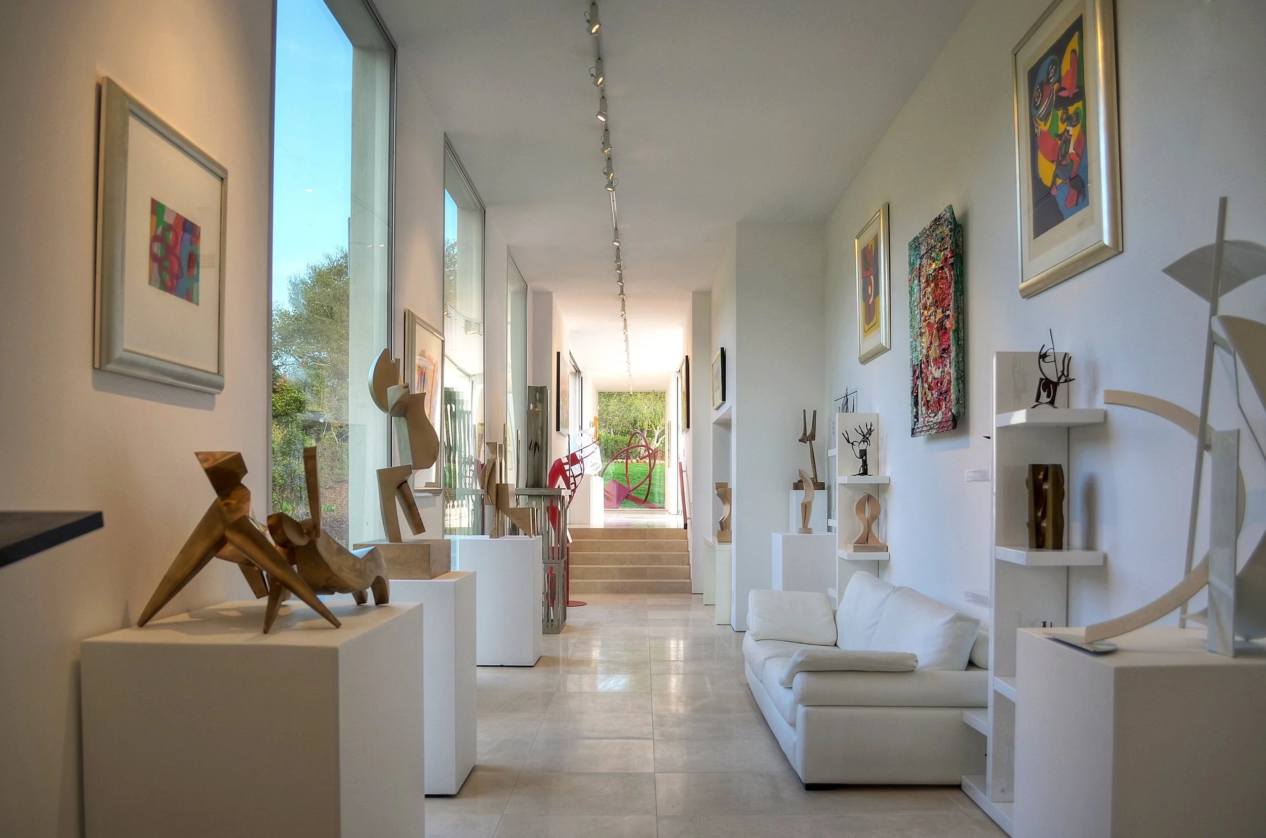 A contemporary art gallery hallway with sculptures and paintings, featuring large windows allowing natural light to enhance the artwork.