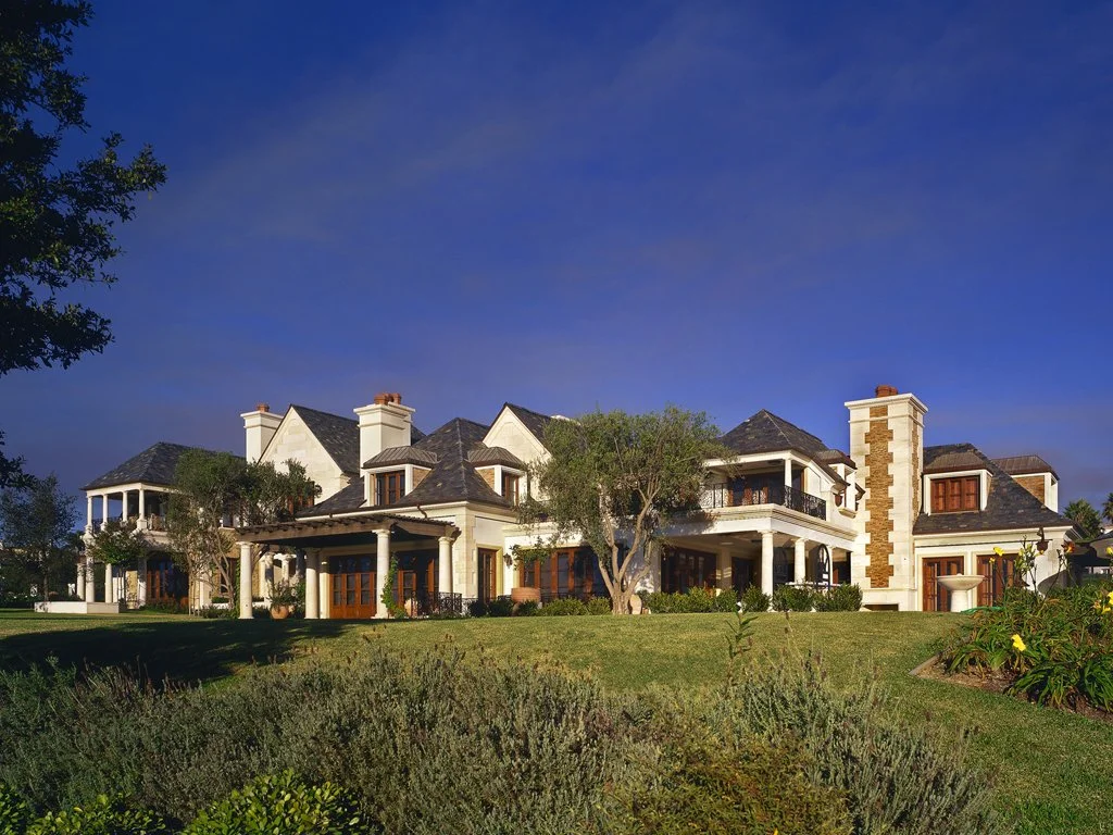 Large luxurious mansion with stone facade, multiple chimneys, and lush landscaping under a clear blue sky.