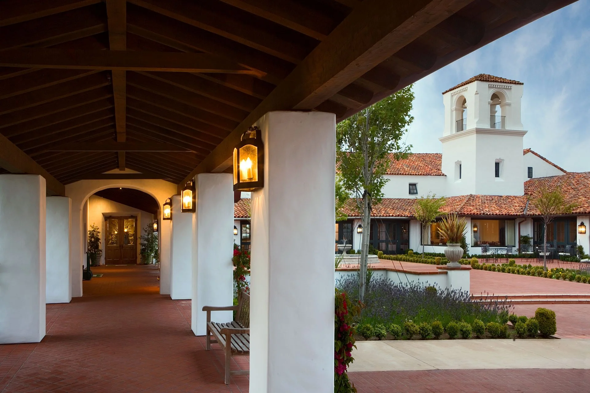 Covered outdoor walkway with lantern-style lighting leading to a courtyard at Monterey Peninsula Country Club, featuring Spanish-style architecture with white stucco walls, terracotta roof tiles, and a bell tower.