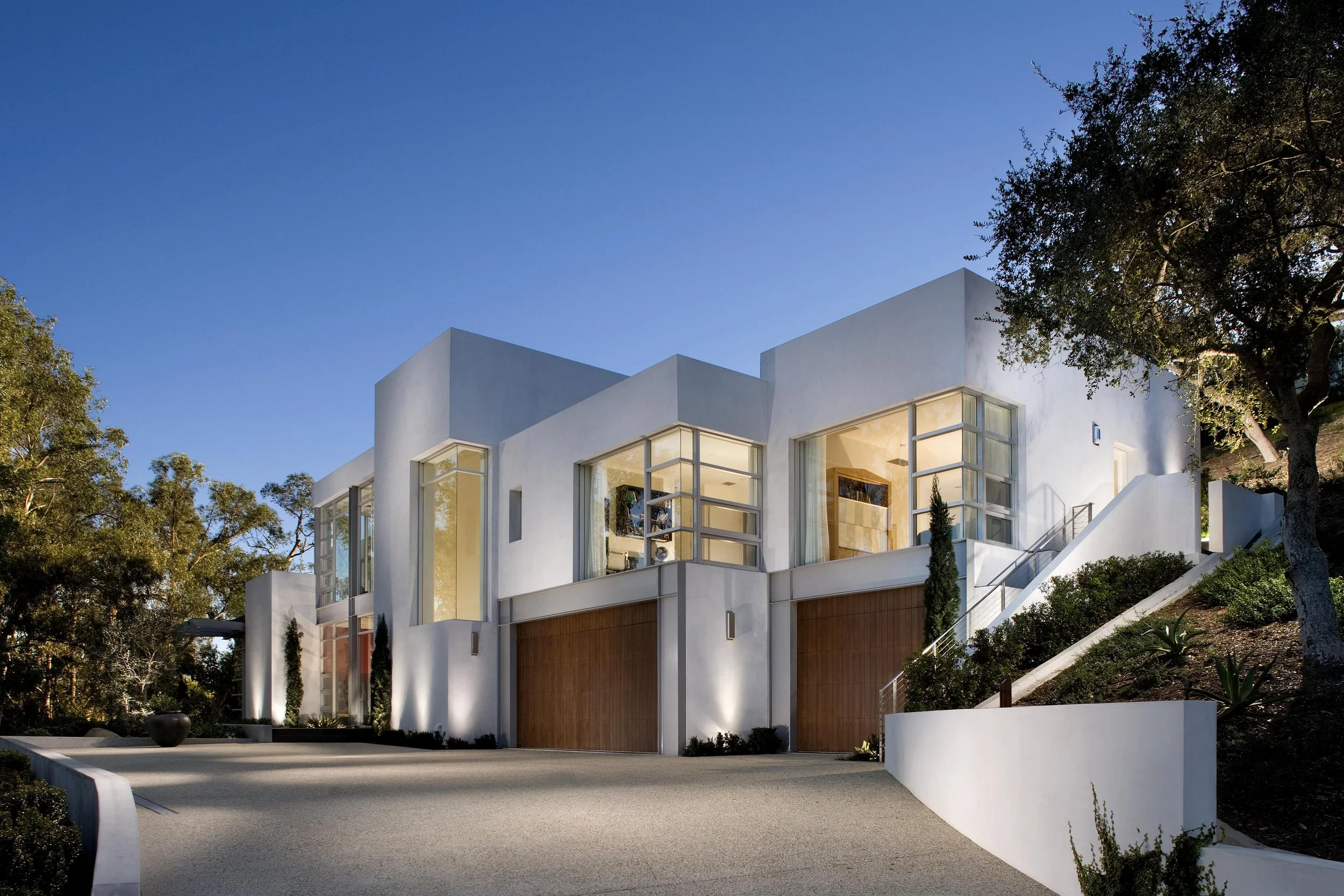 Modern white house with large windows, wooden garage doors, and landscaped driveway, surrounded by trees.