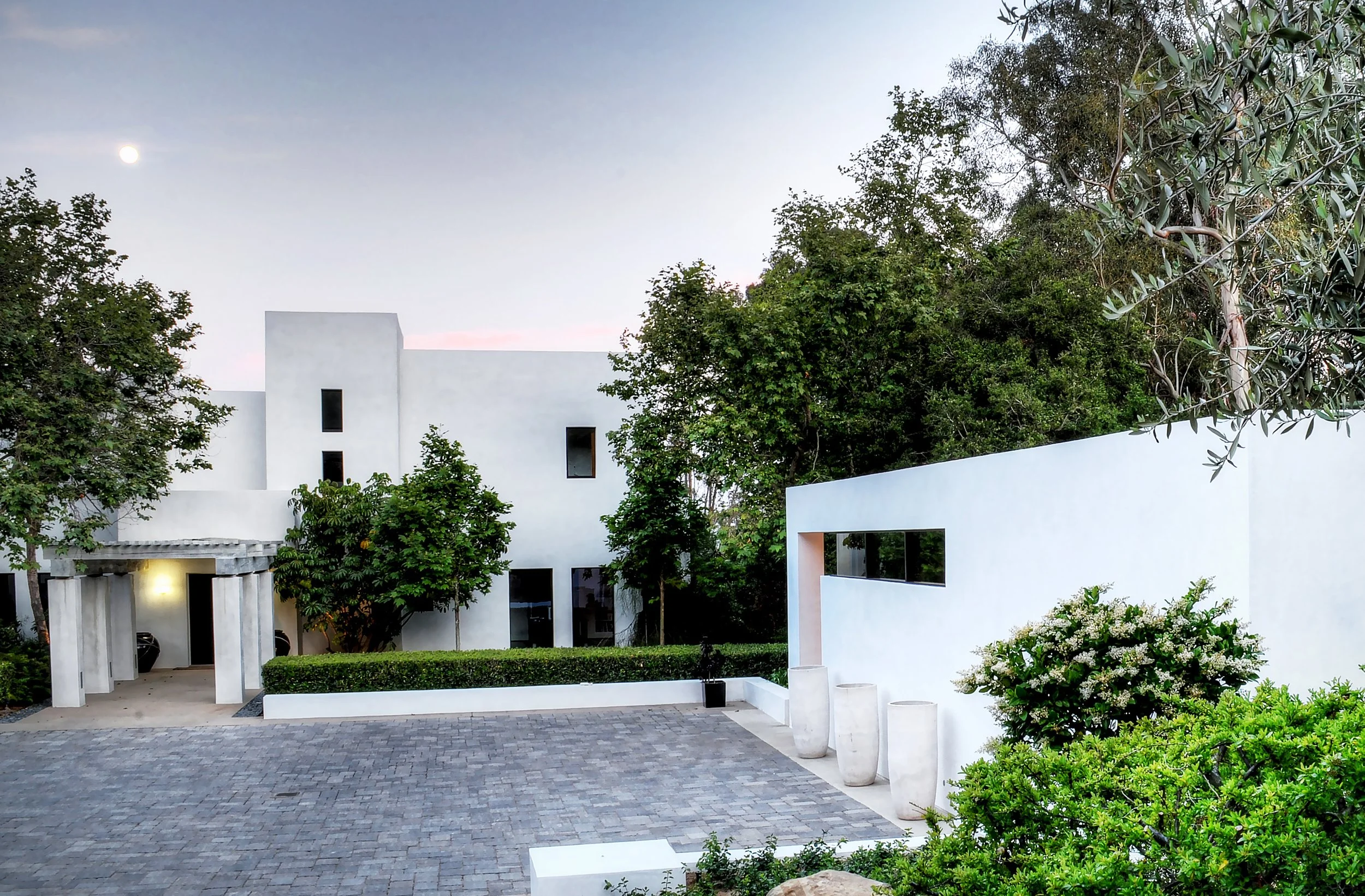 Contemporary white home with minimalist architecture, surrounded by greenery and featuring a paved driveway, designed by The Warner Group Architects, Inc.