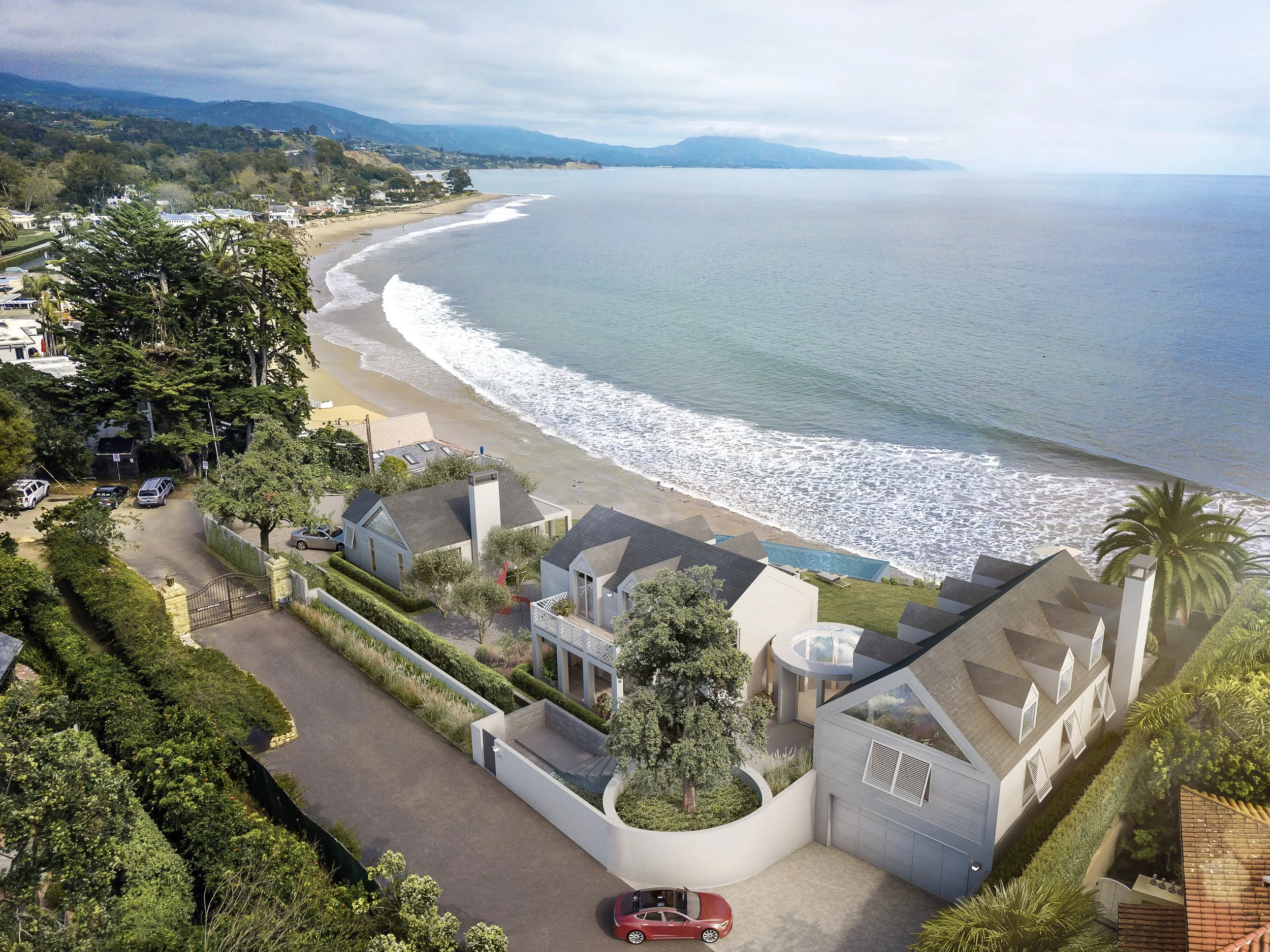 Aerial view of a coastal residential area with modern houses, lush greenery, and a beach along the shoreline, with waves gently lapping against the sand. The background features mountains and a cloudy sky.