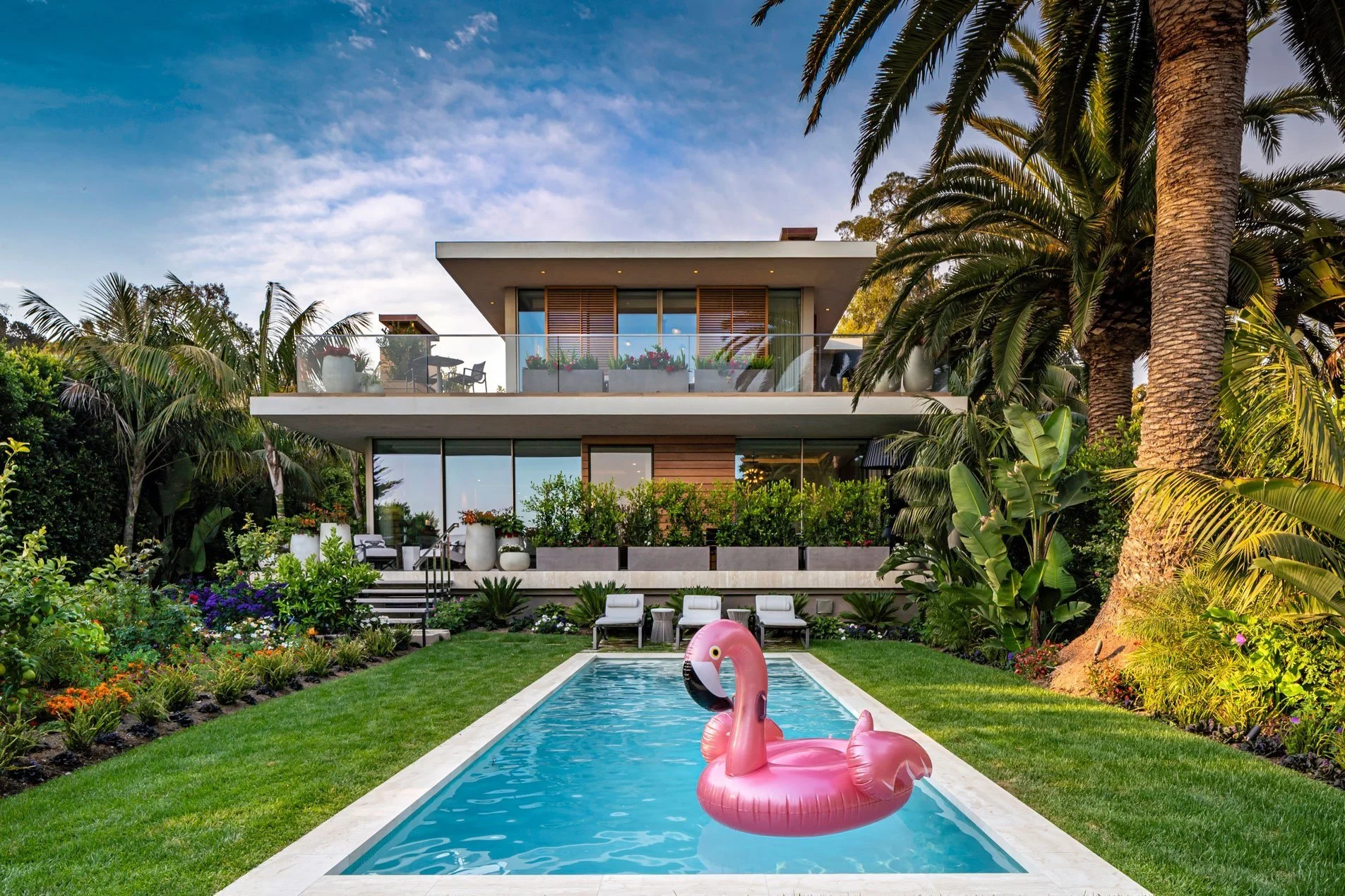 Modern house with glass balcony, lush garden, swimming pool with pink flamingo float, and palm trees.