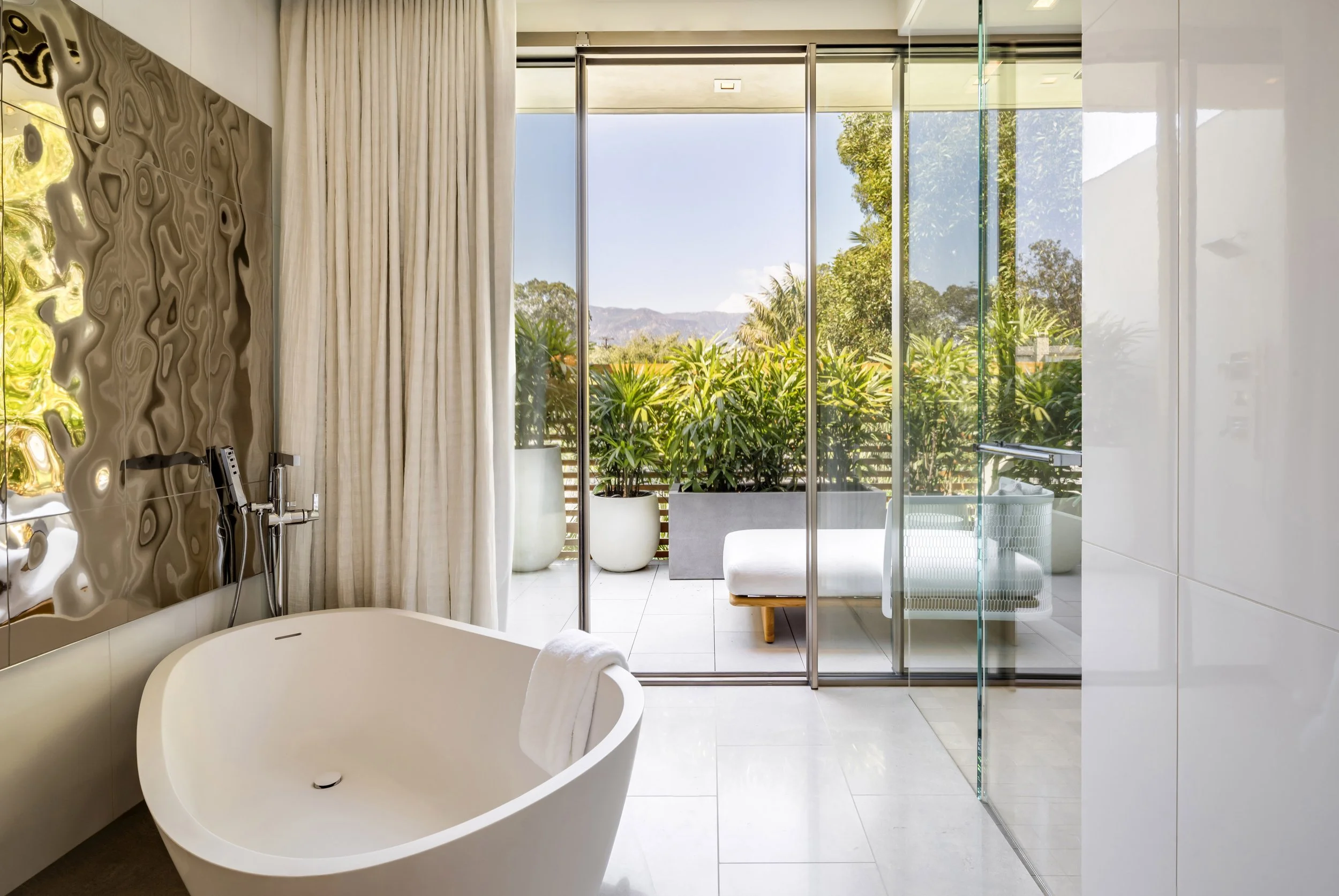 Modern bathroom with freestanding tub, glass shower, large window with garden view, and lounge chair outside.