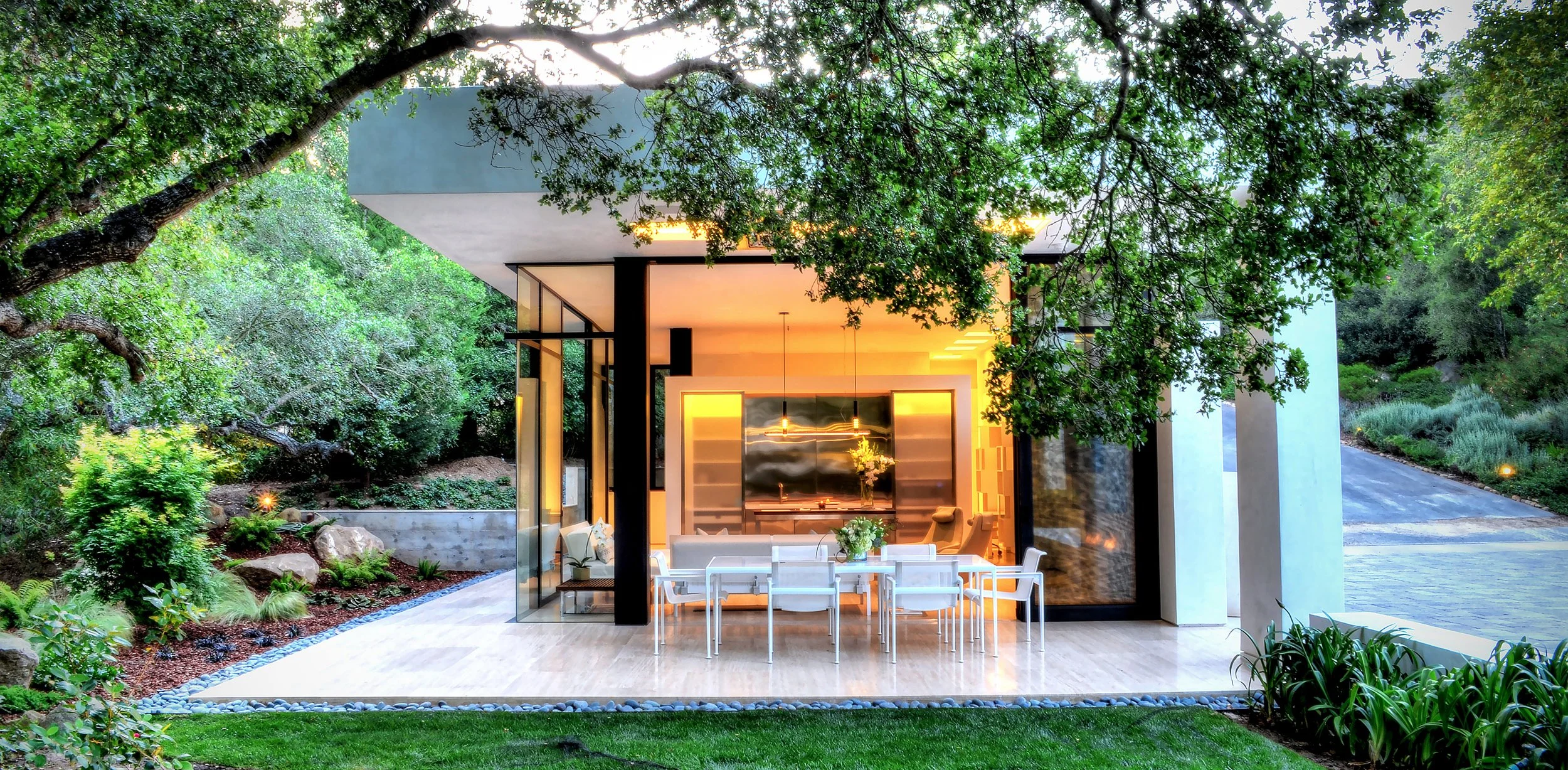 Modern glass-walled house with outdoor dining area surrounded by lush greenery and trees.